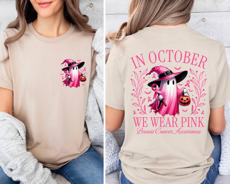 May include: A tan t-shirt with a pink ghost wearing a witch's hat and holding a pumpkin. The back of the shirt says "In October We Wear Pink Breast Cancer Awareness" with a pink ghost wearing a witch's hat and holding a pumpkin.