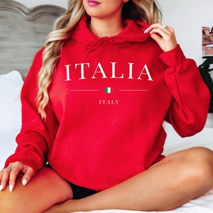 May include: A red hoodie with the word "ITALIA" in white capital letters. Below the word is a horizontal line, a small Italian flag, and the word "ITALY". The hoodie has a drawstring hood and a front pocket.