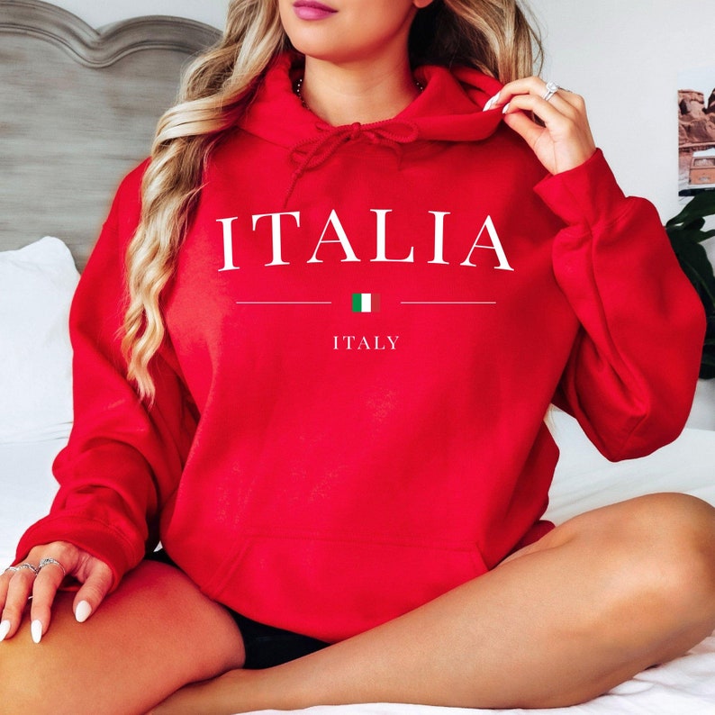 May include: A red hoodie with the word "ITALIA" in white capital letters. Below the word is a horizontal line, a small Italian flag, and the word "ITALY". The hoodie has a drawstring hood and a front pocket.