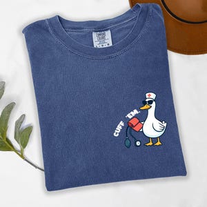 K&ouml;nnte beinhalten: Marineblaues Comfort Colors T-Shirt mit einer Cartoon-Ente, die eine Sonnenbrille und eine Krankenschwesterm&uuml;tze tr&auml;gt und eine Blutdruckmanschette h&auml;lt. Der Text "CUFF 'EM" ist auf dem Shirt aufgedruckt.