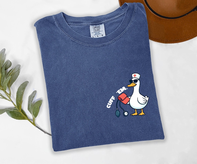 K&ouml;nnte beinhalten: Marineblaues Comfort Colors T-Shirt mit einer Cartoon-Ente, die eine Sonnenbrille und eine Krankenschwesterm&uuml;tze tr&auml;gt und eine Blutdruckmanschette h&auml;lt. Der Text "CUFF 'EM" ist auf dem Shirt aufgedruckt.