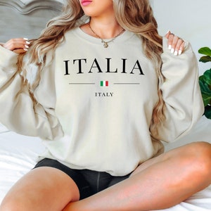 May include: Cream-colored sweatshirt with the word "ITALIA" in black, above a horizontal line and the Italian flag. Below the line, the word "ITALY" is printed in black. The sweatshirt is a casual style.