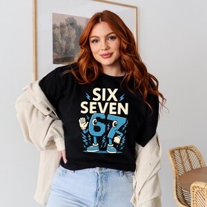 May include: Black t-shirt with the words "SIX SEVEN" in a bold, retro font. The numbers "6" and "7" are designed as cartoon characters in blue, with arms, legs, and shoes. The shirt is worn by a person.