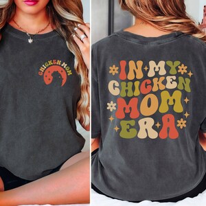 May include: A dark gray t-shirt with a retro-style graphic that says "In My Chicken Mom Era" in colorful letters with stars. The graphic is on the back of the shirt.
