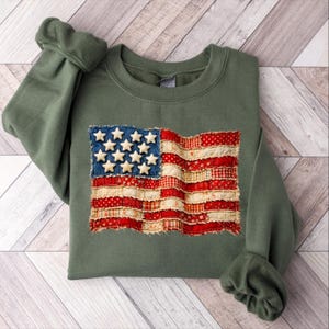 May include: An olive green sweatshirt featuring a patchwork American flag design. The flag has a blue denim field with white stars and red and white striped sections. The sleeves are folded, and the sweatshirt is laid flat.