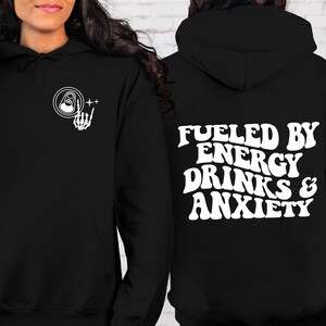 Puede incluir: Sudadera con capucha negra con un gr&aacute;fico blanco de una lata y una mano de esqueleto en la parte delantera y el texto "Fueled by Energy Drinks & Anxiety" en la parte posterior.