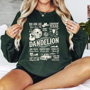 Peut inclure: Un sweat-shirt à manches longues vert foncé avec une impression graphique couleur crème représentant une guitare, un chapeau de cowboy, un camion et un paysage désertique. Le texte inclut des phrases comme You Look Like You Love Me.