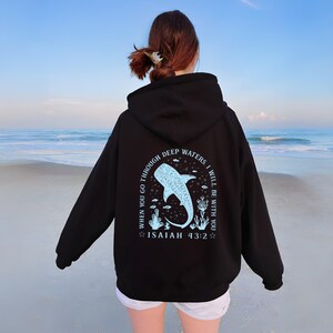 May include: Black hoodie with a light blue whale shark graphic and the text "WHEN YOU GO THROUGH DEEP WATERS I WILL BE WITH YOU" and "ISAIAH 43:2". The hoodie is a pullover style with a hood and long sleeves.