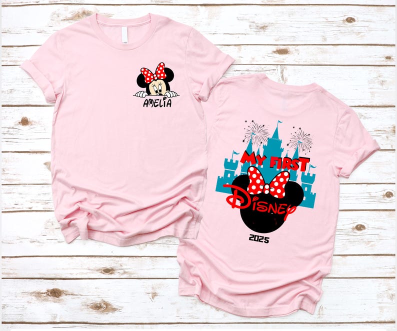 May include: A light pink t-shirt featuring Minnie Mouse. The front of the shirt has Minnie's face with a red bow and the name "Amelia". The back of the shirt has the text "My First Disney" with a castle graphic and the year 2025.