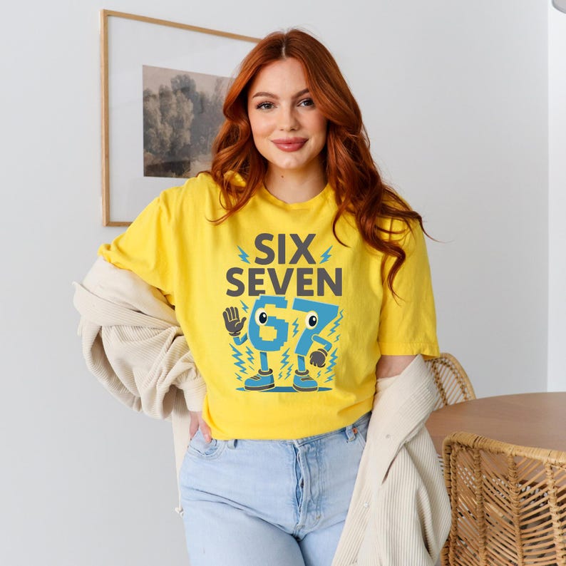 May include: A yellow t-shirt with the text "SIX SEVEN" and a cartoon graphic of the numbers 6 and 7. The numbers have eyes, feet, and are surrounded by lightning bolts. The person is wearing light blue jeans and a beige jacket.