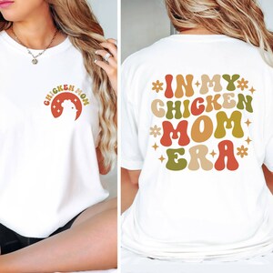 May include: White t-shirt with a retro-style graphic that says "Chicken Mom" on the front and "In My Chicken Mom Era" on the back.