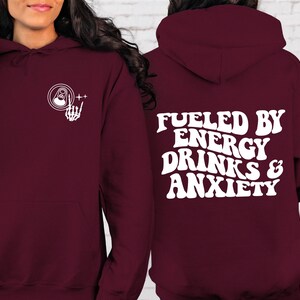 Puede incluir: Sudadera con capucha color burdeos con un gr&aacute;fico blanco de una lata y una mano de esqueleto en el lado izquierdo y el texto "Fueled by Energy Drinks & Anxiety" en una fuente retro en el lado derecho.