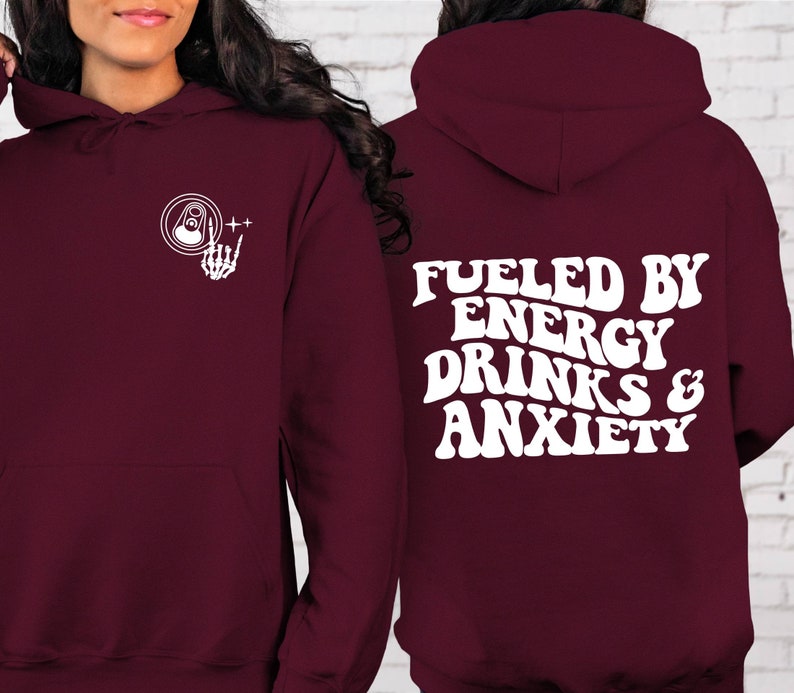 Puede incluir: Sudadera con capucha color burdeos con un gr&aacute;fico blanco de una lata y una mano de esqueleto en el lado izquierdo y el texto "Fueled by Energy Drinks & Anxiety" en una fuente retro en el lado derecho.