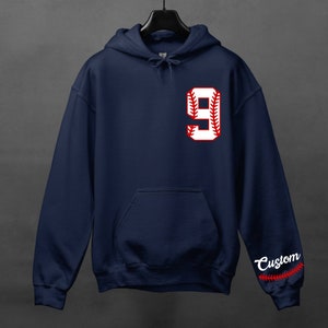 K&ouml;nnte beinhalten: Dunkelblauer Kapuzenpullover mit einer wei&szlig;en Baseball-Nummer 9 auf der Vorderseite. Die Nummer hat rote N&auml;hte. Der linke &Auml;rmel hat das Wort "Custom" in Wei&szlig; mit roten N&auml;hten.