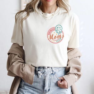 May include: A white t-shirt with a graphic of a melting ice cream cone with a face and the text "Overstimulated Moms Club".