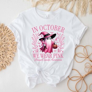 May include: White t-shirt with a pink graphic that reads "IN OCTOBER WE WEAR PINK Breast Cancer Awareness." The graphic features a ghost wearing a witch hat and holding a trick-or-treat pumpkin. The design includes pink ribbons and floral elements.