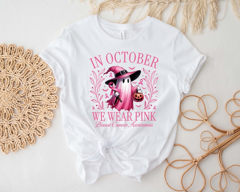 May include: White t-shirt with a pink graphic that reads "IN OCTOBER WE WEAR PINK Breast Cancer Awareness." The graphic features a ghost wearing a witch hat and holding a trick-or-treat pumpkin. The design includes pink ribbons and floral elements.