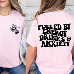 May include: Pink t-shirt with a black graphic on the front of a can with a skeleton hand giving a rock and roll sign. The back of the shirt has the text "Fueled by Energy Drinks & Anxiety" in black.
