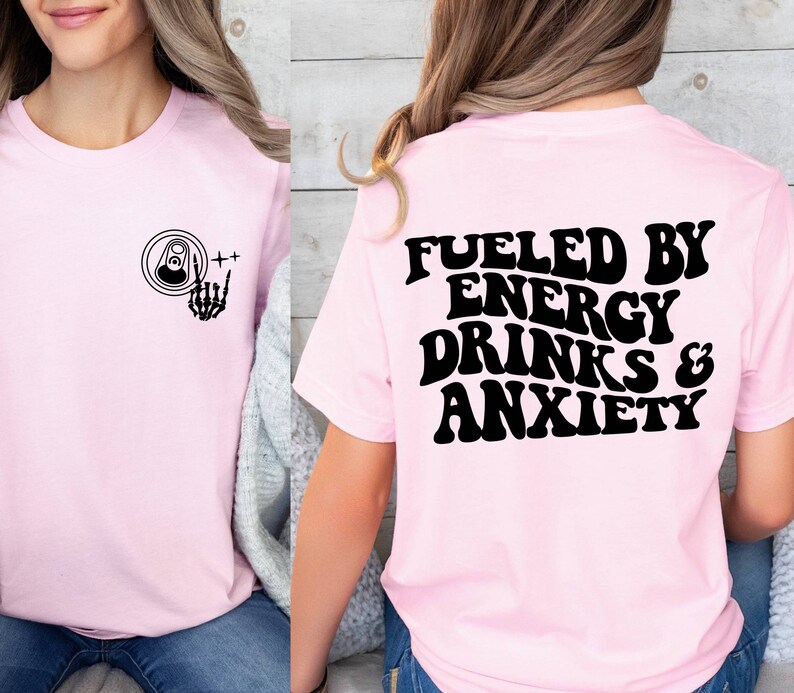 May include: Pink t-shirt with a black graphic on the front of a can with a skeleton hand giving a rock and roll sign. The back of the shirt has the text "Fueled by Energy Drinks & Anxiety" in black.