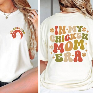 May include: White t-shirt with the text "Chicken Mom Era" in colorful retro font. The text is surrounded by stars and flowers.
