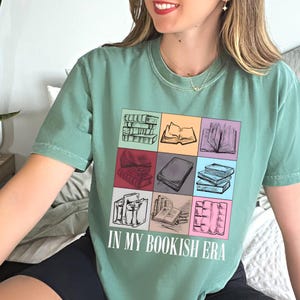 K&ouml;nnte beinhalten: Ein salbeigr&uuml;nes T-Shirt mit einer bunten Grafik von B&uuml;chern in verschiedenen Anordnungen. Der Schriftzug "IN MY BOOKISH ERA" ist darunter in Wei&szlig; aufgedruckt. Das Shirt ist ein bequemer, l&auml;ssiger Stil.
