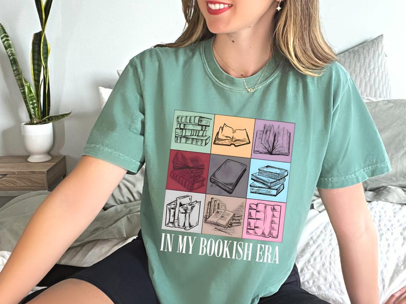 K&ouml;nnte beinhalten: Ein salbeigr&uuml;nes T-Shirt mit einer bunten Grafik von B&uuml;chern in verschiedenen Anordnungen. Der Schriftzug "IN MY BOOKISH ERA" ist darunter in Wei&szlig; aufgedruckt. Das Shirt ist ein bequemer, l&auml;ssiger Stil.