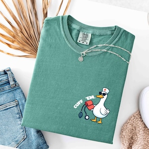 K&ouml;nnte beinhalten: Ein teal-farbenes Comfort Colors T-Shirt mit einer Grafik einer Ente, die einen Krankenschwesterhut und eine Sonnenbrille tr&auml;gt und ein Blutdruckmessger&auml;t h&auml;lt. Der Text "CUFF 'EM." ist auf dem Shirt aufgedruckt. Eine silberne Halskette und eine Jeans sind ebenfalls zu sehen.