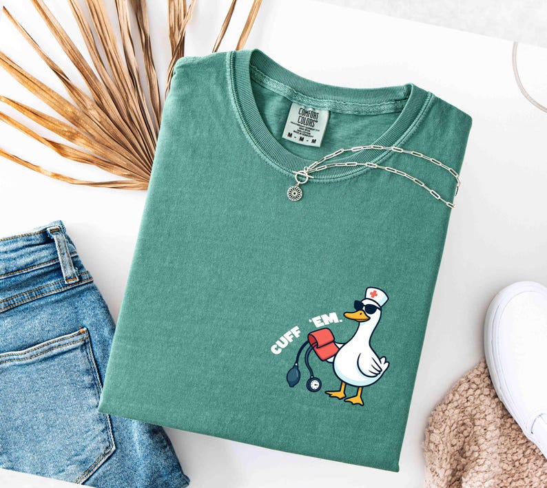 K&ouml;nnte beinhalten: Ein teal-farbenes Comfort Colors T-Shirt mit einer Grafik einer Ente, die einen Krankenschwesterhut und eine Sonnenbrille tr&auml;gt und ein Blutdruckmessger&auml;t h&auml;lt. Der Text "CUFF 'EM." ist auf dem Shirt aufgedruckt. Eine silberne Halskette und eine Jeans sind ebenfalls zu sehen.