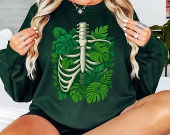 May include: Dark green sweatshirt featuring a white rib cage design filled with green tropical leaves. The design is centered on the front of the sweatshirt. The sweatshirt has long sleeves and a crew neck.