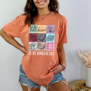 K&ouml;nnte beinhalten: Orangefarbenes T-Shirt mit einer farbenfrohen Grafik von B&uuml;chern in Quadraten und dem Text "IN MY BOOKISH ERA". Das Shirt besteht aus einem weichen Material und hat kurze &Auml;rmel. Das Shirt wird mit Jeansshorts kombiniert.