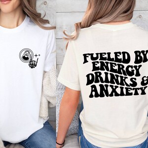 May include: Two white t-shirts with black text and graphics. The front shirt has a skeleton hand holding a can of energy drink. The back shirt says "Fueled by Energy Drinks & Anxiety".