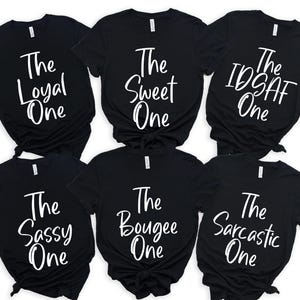 Peut inclure: Six t-shirts noirs avec du texte blanc. Les t-shirts affichent les inscriptions "The Loyal One", "The Sweet One", "The IDSAF One", "The Sassy One", "The Bougee One" et "The Sarcastic One".