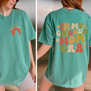 May include: A green t-shirt with the text "In My Chicken Mom Era" printed on the back in colorful, retro-style lettering.