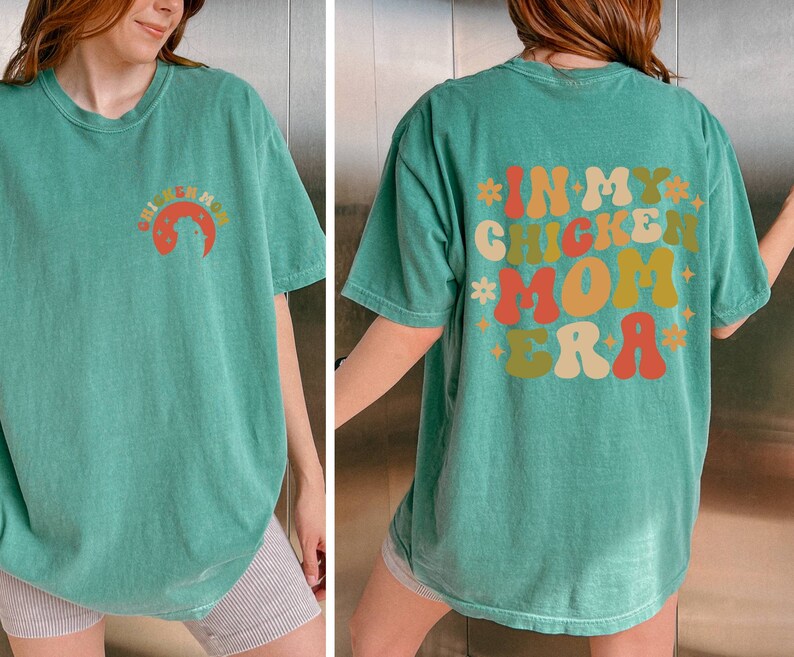 May include: A green t-shirt with the text "In My Chicken Mom Era" printed on the back in colorful, retro-style lettering.