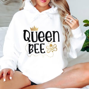 May include: A white hooded sweatshirt with the words "Queen Bee" in black script and a gold crown and bee graphic. The bee graphic includes a dotted line. The sweatshirt is a casual style.