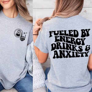 May include: Gray t-shirt with a black graphic on the front of a can with a skeleton hand giving the rock and roll sign. The back of the shirt has the text "Fueled by Energy Drinks & Anxiety" in black.