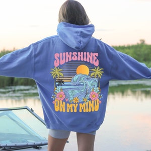 Puede incluir: Una sudadera con capucha azul con un gr&aacute;fico en la espalda que representa una puesta de sol, palmeras y una ola. El texto "SUNSHINE ON MY MIND" est&aacute; impreso encima y debajo de la imagen en rosa y amarillo.
