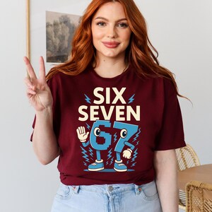 May include: Burgundy t-shirt with the text "SIX SEVEN" and a cartoon graphic of the numbers 67. The numbers are blue with white accents and lightning bolt details. The shirt is a crew neck.