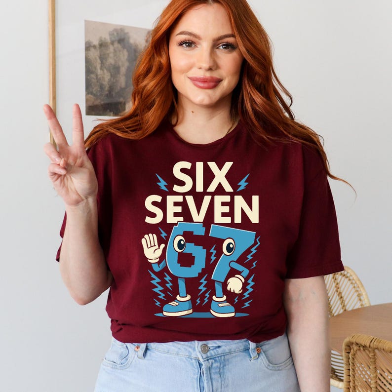 May include: Burgundy t-shirt with the text "SIX SEVEN" and a cartoon graphic of the numbers 67. The numbers are blue with white accents and lightning bolt details. The shirt is a crew neck.