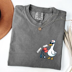 K&ouml;nnte beinhalten: Ein graues Comfort Colors T-Shirt mit einer Grafik einer Ente mit Sonnenbrille und Krankenschwesterm&uuml;tze, die eine Blutdruckmanschette h&auml;lt. Der Text "CUFF 'EM" ist auf dem Shirt aufgedruckt. Eine goldene Halskette liegt &uuml;ber dem Shirt.