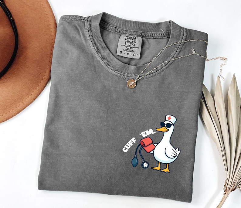 K&ouml;nnte beinhalten: Ein graues Comfort Colors T-Shirt mit einer Grafik einer Ente mit Sonnenbrille und Krankenschwesterm&uuml;tze, die eine Blutdruckmanschette h&auml;lt. Der Text "CUFF 'EM" ist auf dem Shirt aufgedruckt. Eine goldene Halskette liegt &uuml;ber dem Shirt.