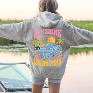 Puede incluir: Una sudadera con capucha gris claro con un gr&aacute;fico colorido en la espalda. El gr&aacute;fico presenta las palabras "SUNSHINE ON MY MIND" en rosa y amarillo, junto con una puesta de sol, palmeras y una ola con flores. La sudadera tiene una capucha con cord&oacute;n.