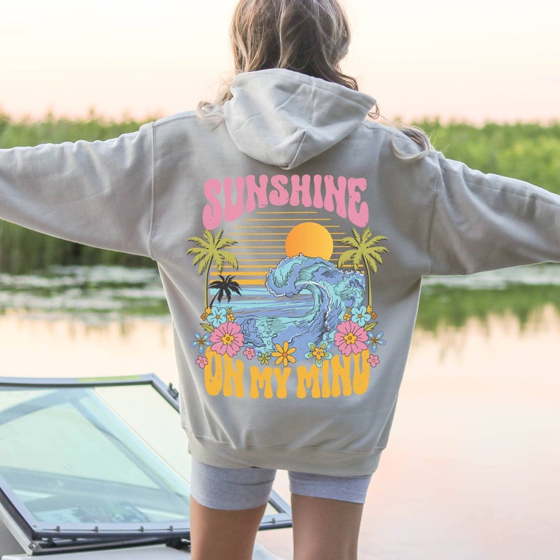 Puede incluir: Una sudadera con capucha gris claro con un gr&aacute;fico colorido en la espalda. El gr&aacute;fico presenta las palabras "SUNSHINE ON MY MIND" en rosa y amarillo, junto con una puesta de sol, palmeras y una ola con flores. La sudadera tiene una capucha con cord&oacute;n.
