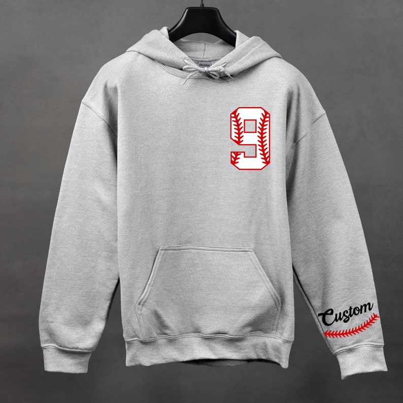 K&ouml;nnte beinhalten: Ein grauer Kapuzenpullover mit einer wei&szlig;en Baseballnummer 9 auf der Vorderseite und dem Wort "Custom" mit einem Baseball-Stichdesign auf dem &Auml;rmel.