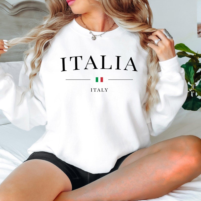 May include: White sweatshirt with the word "ITALIA" in black capital letters. Below the word is a horizontal line, the Italian flag, and the word "ITALY". The sweatshirt is a crew neck style.