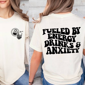 May include: White t-shirt with a black graphic on the front of a can with a skeleton hand giving a rock and roll sign. The back of the shirt has the text "Fueled by Energy Drinks & Anxiety" in black.