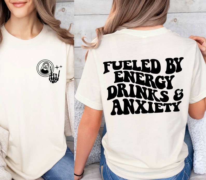 May include: White t-shirt with a black graphic on the front of a can with a skeleton hand giving a rock and roll sign. The back of the shirt has the text "Fueled by Energy Drinks & Anxiety" in black.