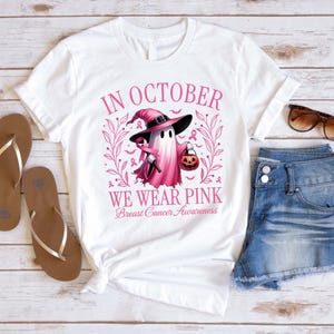 May include: White t-shirt with a pink graphic featuring a ghost wearing a witch hat and holding a pumpkin. The text "IN OCTOBER WE WEAR PINK Breast Cancer Awareness" is printed on the shirt. Brown sandals and denim shorts are also visible.