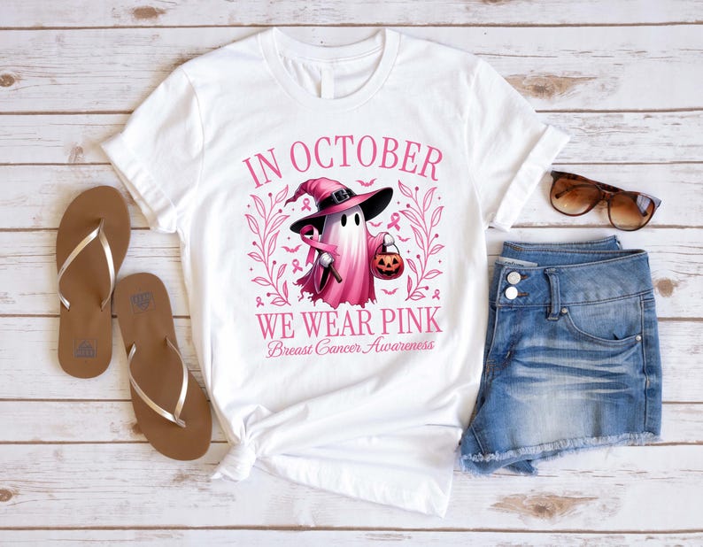 May include: White t-shirt with a pink graphic featuring a ghost wearing a witch hat and holding a pumpkin. The text "IN OCTOBER WE WEAR PINK Breast Cancer Awareness" is printed on the shirt. Brown sandals and denim shorts are also visible.
