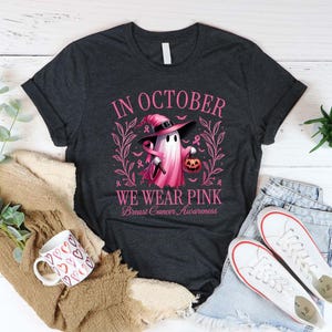May include: Dark gray t-shirt with the text "IN OCTOBER WE WEAR PINK Breast Cancer Awareness". The shirt features a pink ghost wearing a witch hat and holding a pumpkin. A coffee mug with heart designs and white sneakers are also visible.